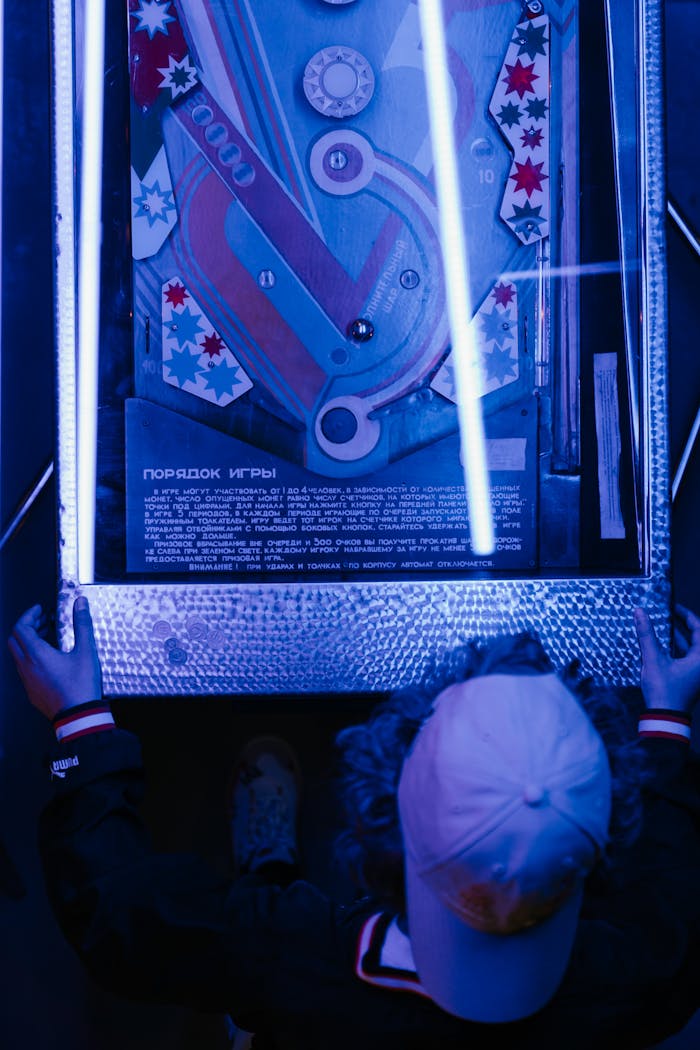 Teenager in a cap playing retro pinball machine under neon lights for a nostalgic arcade vibe.